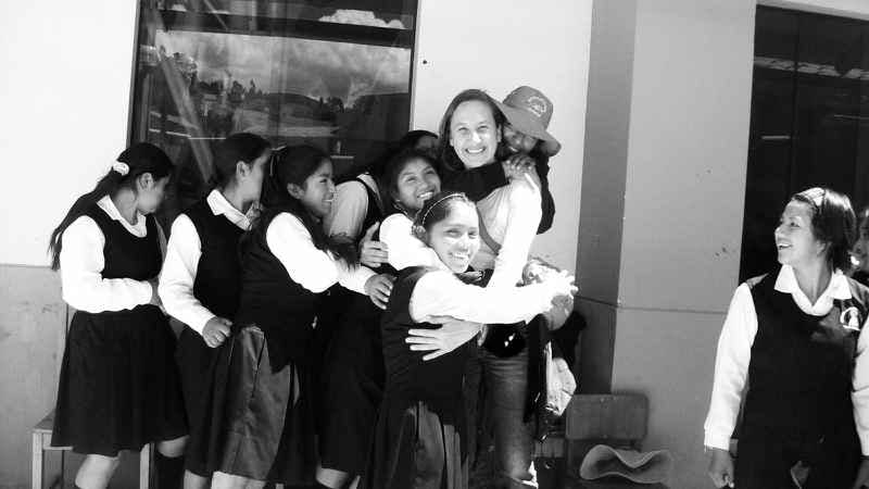 Nuria Gracia, center, embraces a group of seven girls wearing uniforms during her time in Peru. One girl looks on. The photo is taken outside of a building and is black and white.